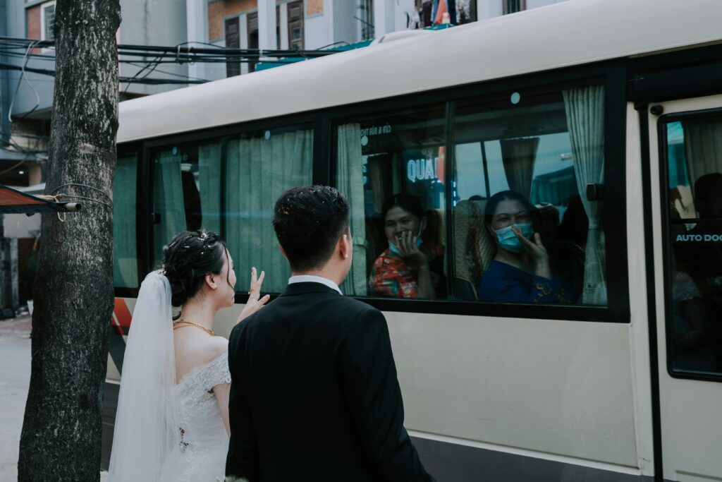 Young bride and groom saying goodbye and waving hands to guests in masks sitting in bus
